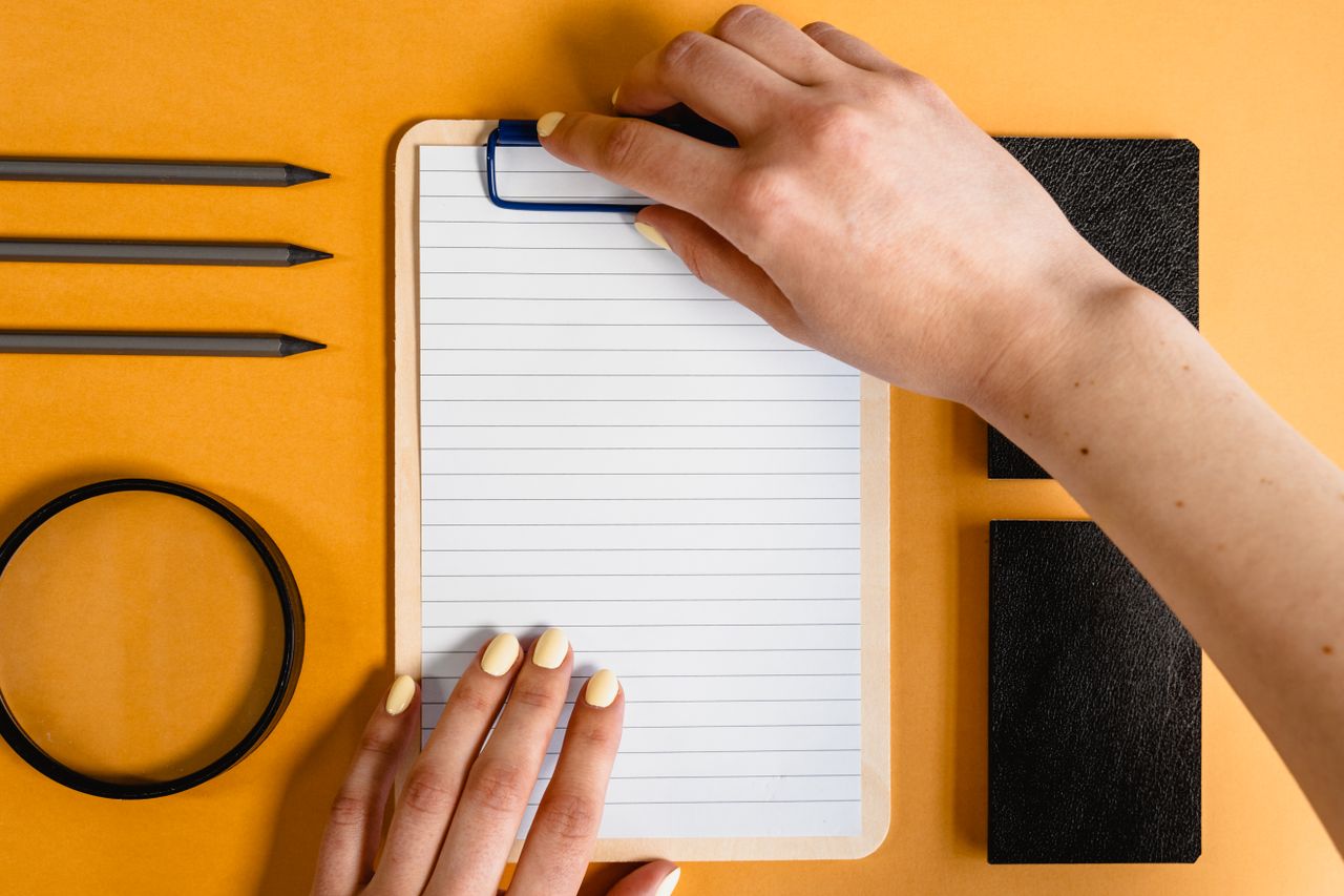 Top view of hands on a notepad with various stationery items on a yellow desk background.