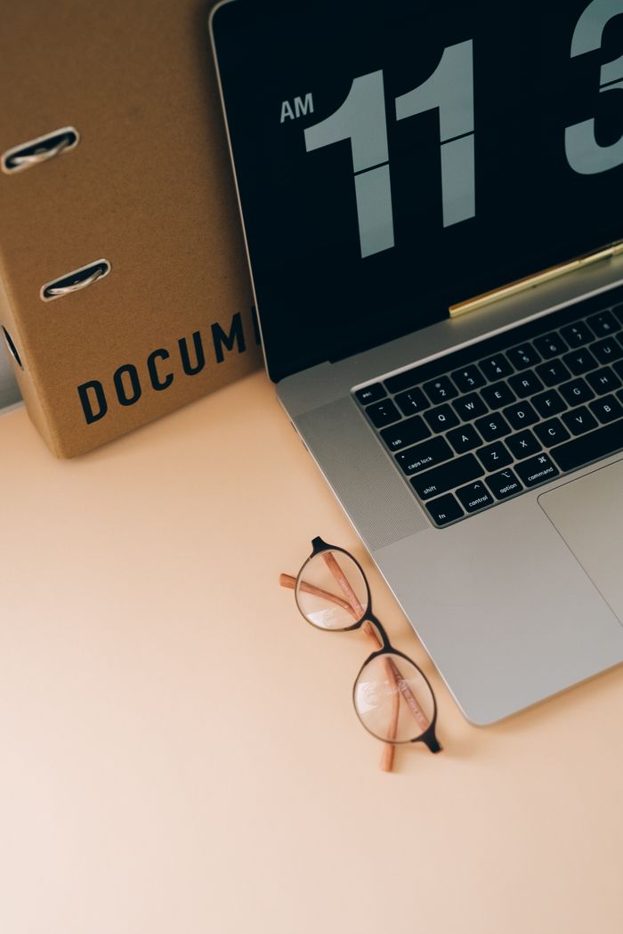 Flat lay of a laptop, eyeglasses, and document binder on a beige desk, perfect for workspace concepts.